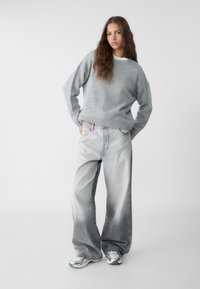 Helles graues Strickpullover mit ribbeligem Saum, kombiniert mit übergroßen, verblassten grauen Jeans und silbernen Sneakers, vor einem schlichten Hintergrund.