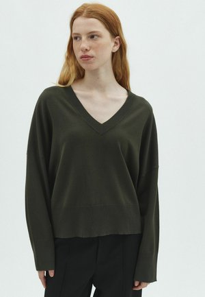 Jeune personne aux longs cheveux roux portant un pull vert foncé à col en V et un pantalon noir, regardant légèrement de côté sur un fond uni.