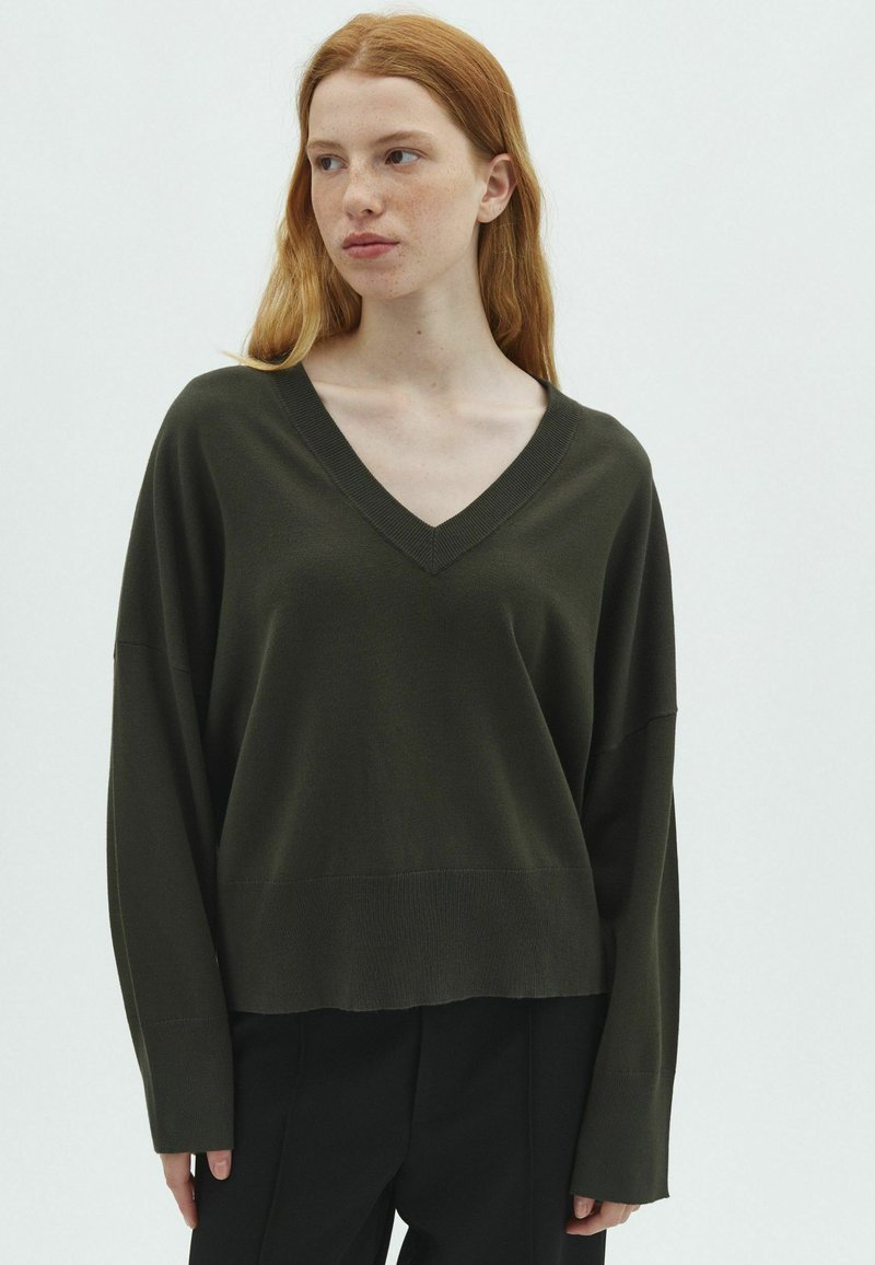 Jeune personne aux longs cheveux roux portant un pull vert foncé à col en V et un pantalon noir, regardant légèrement de côté sur un fond uni.