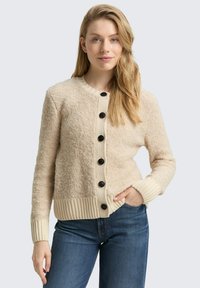 Beige, strukturierter Cardigan mit rundem Ausschnitt, gerippten Bündchen und einer Knopfleiste mit fünf schwarzen Knöpfen. Kombiniert mit blauen Jeans.