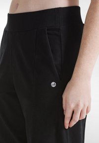 Schwarze lockere Hose mit breitem Bündchen, seitlichen Taschen und einem runden weißen Logo am linken Bein. Weicher Stoff.