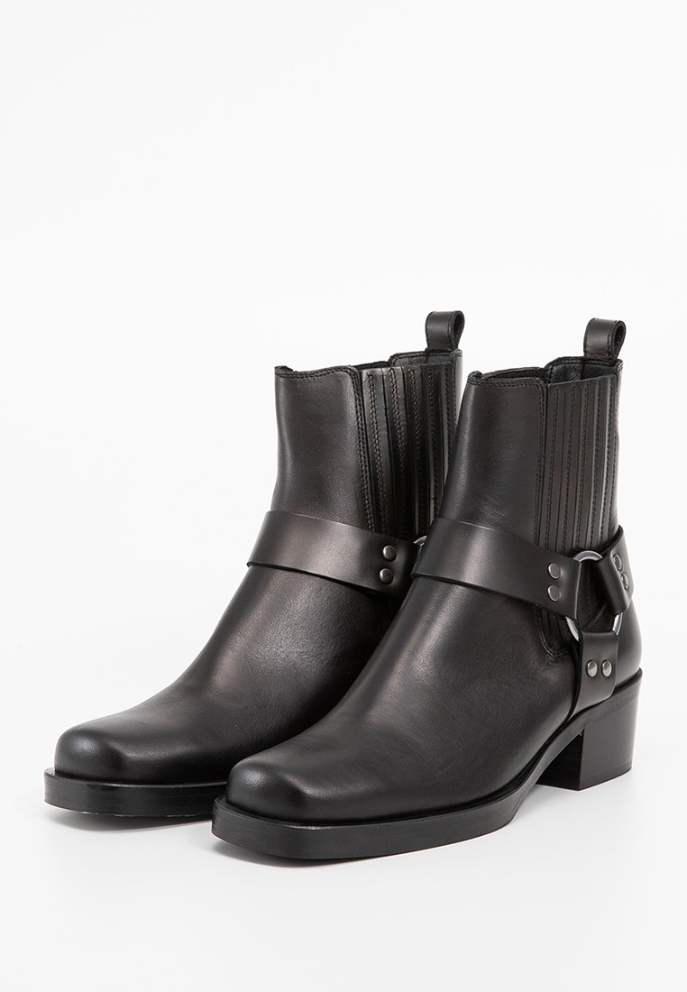 Stivali alla caviglia in pelle nera con punta arrotondata, dotati di pannelli laterali a coste e una larga patta con dettagli in metallo. Design con tacco a blocco.