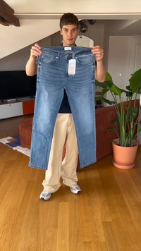 Jovem a stander dentro de casa segurando um par de jeans azuis com etiquetas à frente de um sofá e de uma planta em vaso.