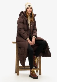 Brun pufferjakke med hætte, beige sweater, sorte leggings og brune snørebåndsstøvler med gule snørebånd; siddende på en træstol.