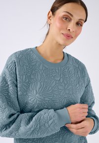 Sudadera acolchada de color azul claro con estampado floral, cuello redondo y puños acanalados. Textura suave, con diseños tridimensionales prominentes.