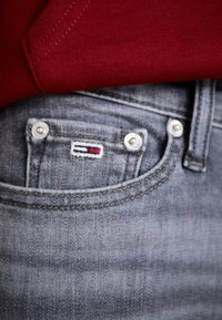 Jeans de mezclilla grises con costuras visibles, herrajes metálicos y una pequeña etiqueta de tela con rayas rojas y azules en el bolsillo delantero.