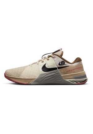 Baskets basses Nike beige et marron clair avec un swoosh noir, une partie avant en mesh à motif, une semelle rembourrée et des accents rouges sur la semelle extérieure et les coutures.