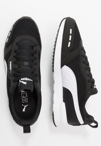 Pantofi sport negri, cu un superior din plasă, accente din piele întoarsă, un logo alb și o talpă texturată. Încălțările includ șireturi și o curea pentru călcâi.