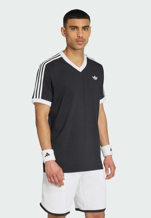 Młody mężczyzna w czarnej koszulce Adidas z dekoltem w serek z białymi paskami na rękawach, białych szortach i białych opaskach na nadgarstki stoi na jednolitym tle.