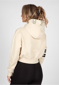 Felpa corta color crema con cappuccio regolabile, polsini a costine e dettagli di branding neri sul retro, abbinata a leggings neri a vita alta.