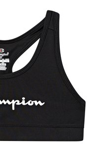 Melna čempiona sporta krūšturis ar racerback dizainu un balto rakstveida logo priekšpusē, redzamu iekšējo birku ar izmēru un ražošanas detaļām.