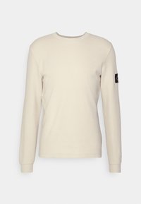 Beige long-sleeve shirt made from a textured fabric. Features a round neckline and a black logo patch on the left sleeve.
