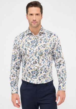 Mann trägt ein weißes Langarmhemd mit blauem, gelbem und grünem floralen Paisleymuster, kombiniert mit dunkelblauen Hosen, und steht vor einem weißen Hintergrund.