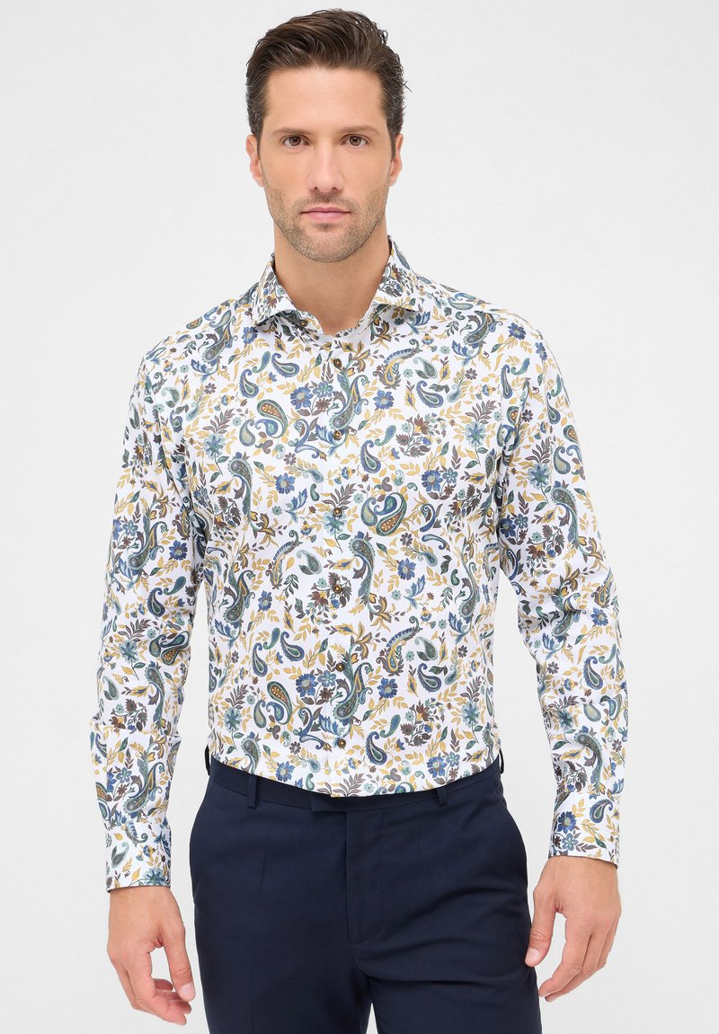 Man in een witte lange mouw shirt met een blauw, geel en groen bloemig paisley patroon, in combinatie met donkere blauwe broek, staand tegen een witte achtergrond.