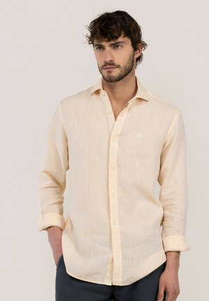 Hombre con cabello rizado oscuro y barba que lleva una camisa beige de manga larga con puños enrollados, de pie con una mano en el bolsillo.
