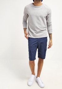 Chemise à manches longues grise, shorts cargo navy avec poches latérales, assortis avec des baskets blanches. Le matériau semble doux ; la chemise a une coupe décontractée.