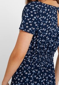 Námořnické květinové šaty s krátkými rukávy, elastickým pasem a světlomodrými a bílými květinovými vzory. Detail ve tvaru klíčové dírky na zadním výstřihu.
