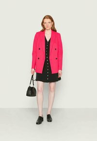 Blazer duplo em rosa choque sobre um vestido preto com botões à frente, combinado com sapatos pretos e uma pequena bolsa preta. Fundo simples.