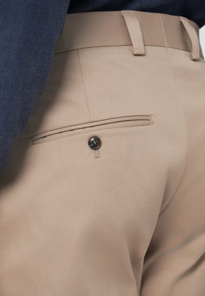 Bēši bikses ar gludu tekstūru, ar vienu aizmugurējo kabatu, kurā ir apaļa melna pogu un jostas cilpas.