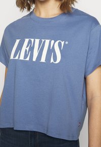 Personne portant un t-shirt court bleu avec un texte blanc "LEVI'S" sur la poitrine et un pantalon foncé.