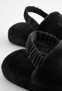Pantofole nere in peluche con una texture morbida, dotate di cinturini in tessuto elastico per una vestibilità sicura e forma arrotondata. Senza hardware visibile.