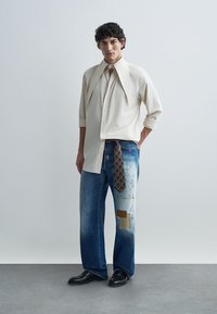 Beige button-up shirt with oversized collar, paired with light blue denim jeans featuring patches and a brown patterned tie at the waist. Black shoes.