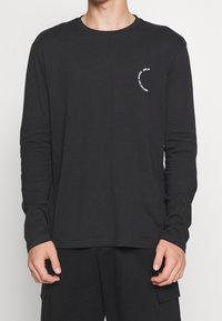 Langärmliges schwarzes T-Shirt aus weicher Baumwolle mit rundem Halsausschnitt und einem weißen runden Logo, das auf der Brust aufgedruckt ist.