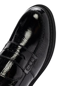 Zapato de cuero negro con un acabado brillante, punta redondeada y una correa decorativa. Costuras notables y una suela de color contrastante.