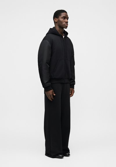 Μαύρο φούτερ με κουκούλα και εμπρόσθια τσέπη, συνδυασμένο με μαύρο παντελόνι φαρδιάς γραμμής και μαύρα παπούτσια· το ύφασμα φαίνεται λείο και παχύ.