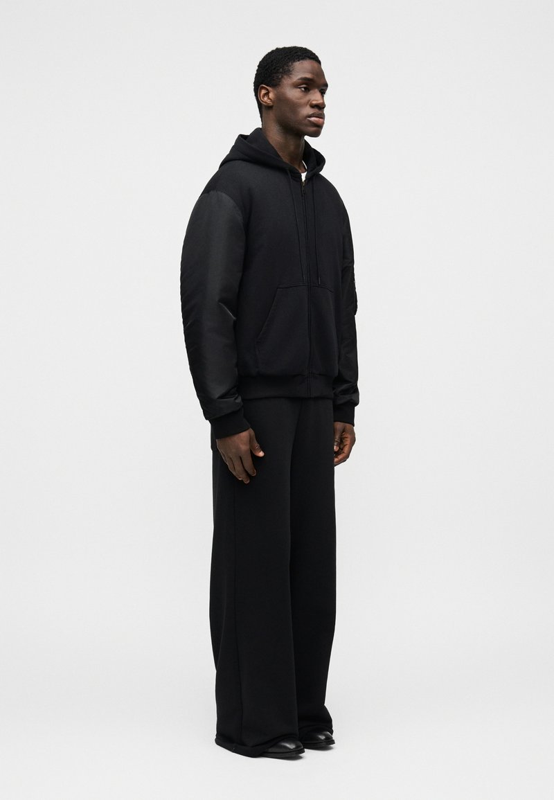 Sort hættetrøje med frontlomme, kombineret med vidt benede sorte bukser og sorte sko; stoffet ser glat og tykt ud.