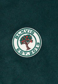 Ümmargune plaastriga, millel on tikkitud õunapuu ja punased õunad, ümber millest on tekst "BLKVIS EST. 2013" tumerohelisel kangal.