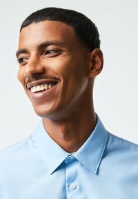 Camicia azzurro chiaro con colletto e chiusura a bottone, caratterizzata da una texture liscia e un design classico, indossata da una persona sorridente.
