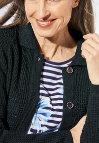 Schwarze strukturierte Strickjacke mit Knopfleiste über einer gestreiften Blumenbluse in Blau- und Weißtönen.