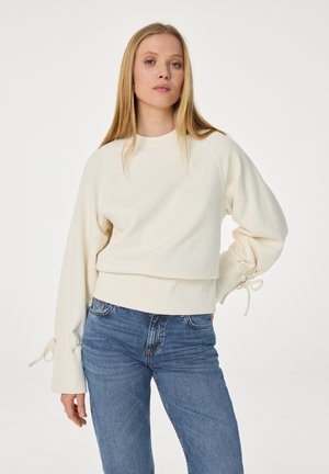 Vrouw met lang blond haar, gekleed in een crèmekleurige trui met lange mouwen en strikjes op de mouwen, en blauwe jeans, staand tegen een effen achtergrond.
