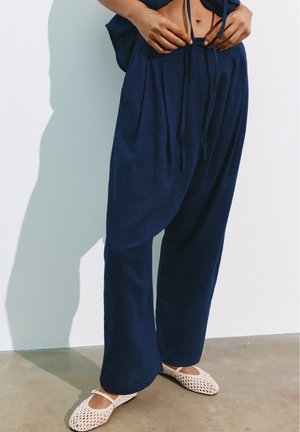Person trägt lockere dunkelblaue Hose und weiße gewebte Ballettschuhe und steht vor einer weißen Wand und einem Betonboden.