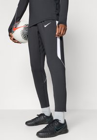 Μαύροι αθλητικοί φόρμες με λευκές πλευρικές λεπτομέρειες, κατασκευασμένες από ελαστικό ύφασμα. Φορεμένοι με μαύρα ποδοσφαιρικά παπούτσια και κρατώντας μια μπάλα ποδοσφαίρου μπροστά.