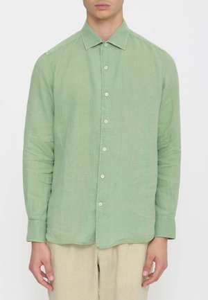 Uomo che indossa una camicia verde chiaro a maniche lunghe con colletto e pantaloni beige, in piedi con le braccia rilassate lungo i fianchi.
