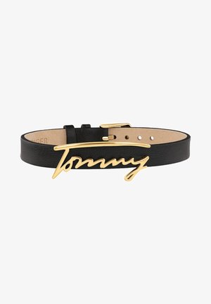 Ceinture en cuir noir avec boucle dorée formant le mot "Tommy" en écriture cursive, avec des trous réglables pour la taille.
