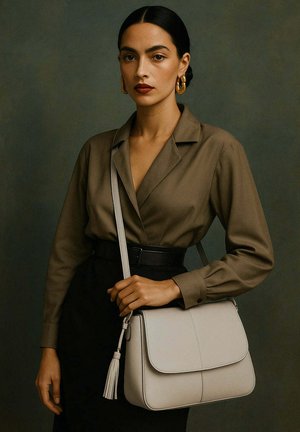 Femme aux cheveux foncés portant un blouse marron et une jupe noire, tenant un sac en bandoulière beige clair avec un pompon contre un fond sombre.