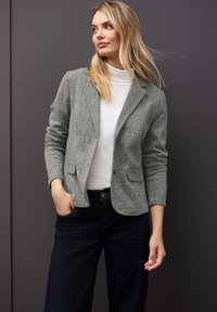 Grå vävd blazer med snedskurna slag, två framfickor och en knappknäppning, i kombination med en vit polotröja och mörka jeans.