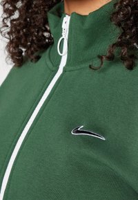 Vihreä vetoketjullinen takki, joka on tehty pehmeästä puuvillasekoitteesta. Takissa on korkea kaulus, valkoinen vetoketju ja pieni musta Nike-logo rinnassa.