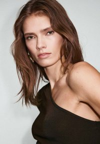 Modelo con cabello largo castaño, vistiendo un top negro de un solo hombro. Su tono de piel es claro, con un maquillaje sutil y pendientes visibles. Fondo neutro.