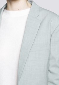 Jasnoszara marynarka z teksturowanej tkaniny, z notched klapą i boczną kieszenią, noszona na białym swetrze z prążkowanym dekoltem.