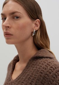 Brauner strukturierter Pullover mit Kragen, silberne Tropfohringe und langes Haar. Der Hintergrund ist neutral und schlicht.