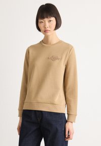 Donna con capelli corti e scuri, che indossa una felpa beige con logo ricamato e jeans blu scuro, in piedi davanti a uno sfondo bianco.