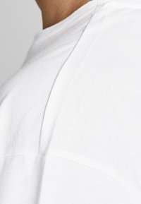 Witte katoenen T-shirt, close-up van de schoudernaden, gladde textuur, ronde kraag en zichtbare stikdetails.