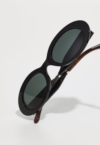 Schwarze übergroße Sonnenbrille mit runden Gläsern, aus glänzendem Kunststoff gefertigt. Die Bügel zeigen ein braunes Schildpatt-Muster und metallische Akzente.