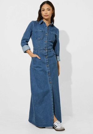 Kvinde iført lang blå denim kjole med knapper og lommer, stående med den ene hånd i lommen og hvide sneakers med sorte striber.