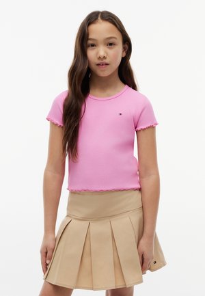 Fille aux longs cheveux bruns portant un t-shirt rose à manches courtes et une jupe plissée beige, debout devant un fond blanc uni.