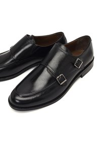 Zapatos de cuero negro con acabado brillante, que cuentan con doble hebilla, puntera redondeada y un tacón bajo apilado. Textura suave y diseño elegante.